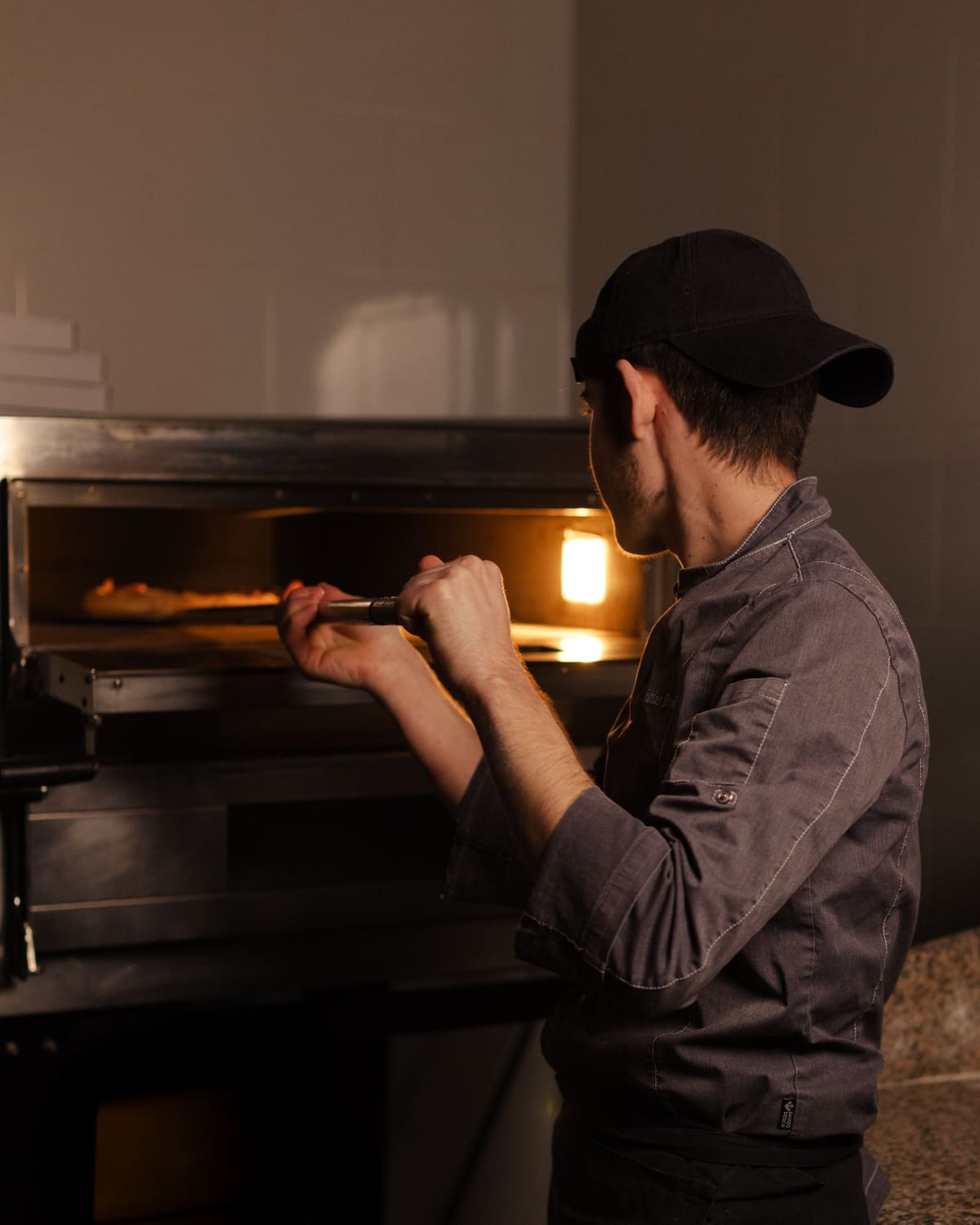 A pizza maker in a cap and grey shirt sliding a pizza into a glowing deck oven with a long metal peel, the oven's warm light on his face.