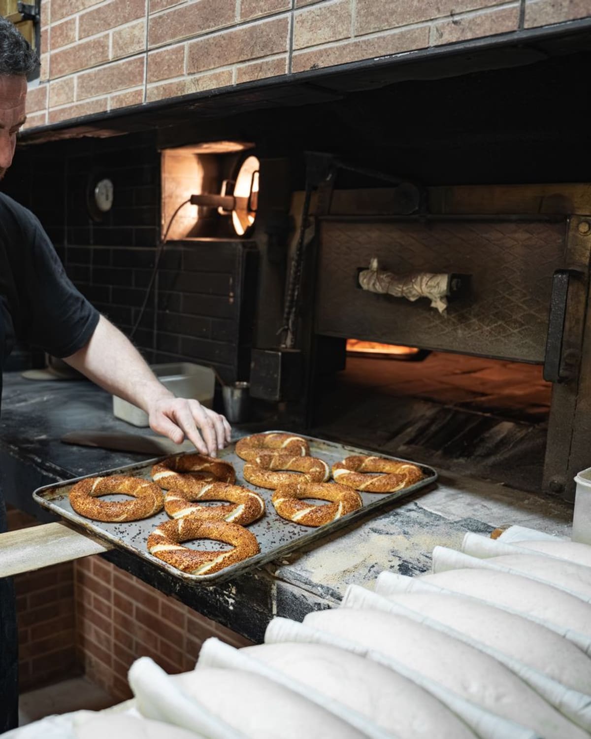 A baker sliding a tray of freshly made pretzels into a brick oven.