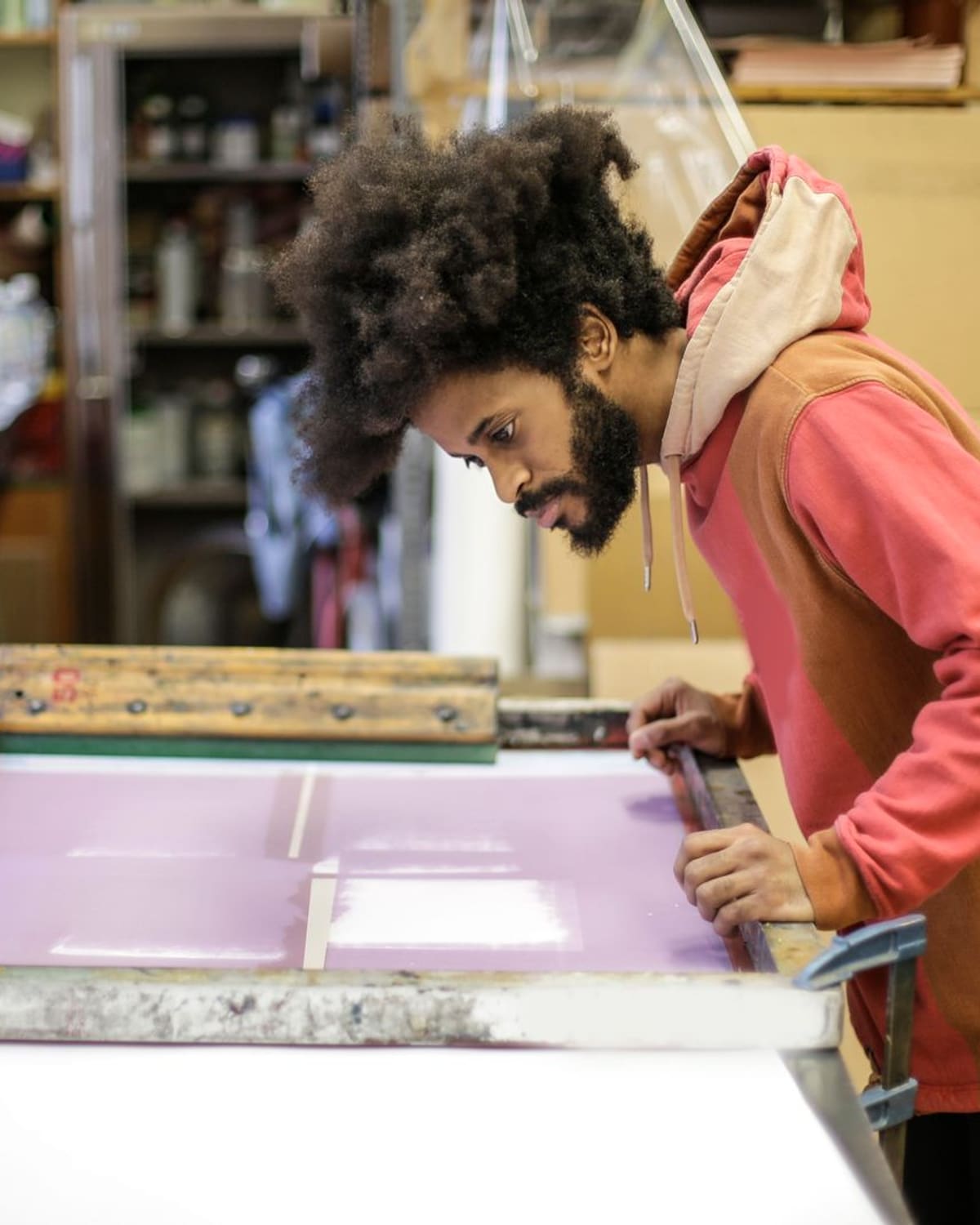 A man leans over a large screen printing frame with purple ink in a workshop, pulling a print.