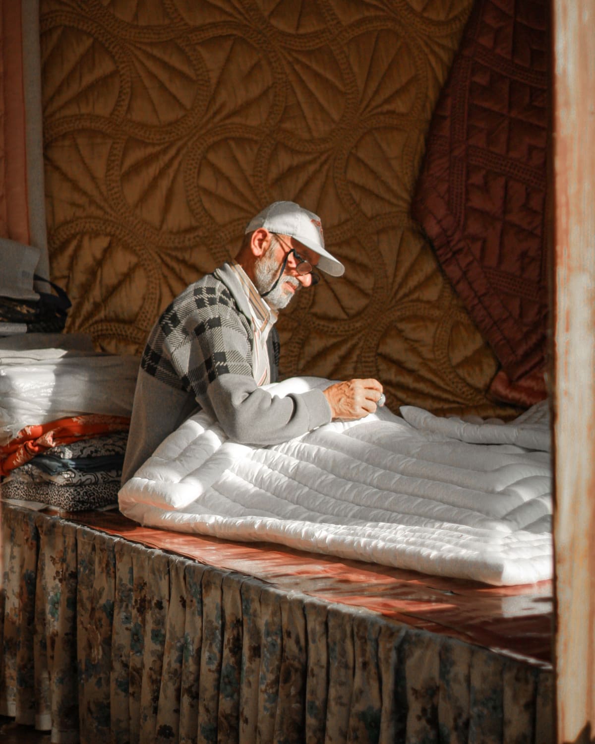 An older man in a cap hand-stitches a white quilt in warm sunlight, finished quilts hanging behind him.
