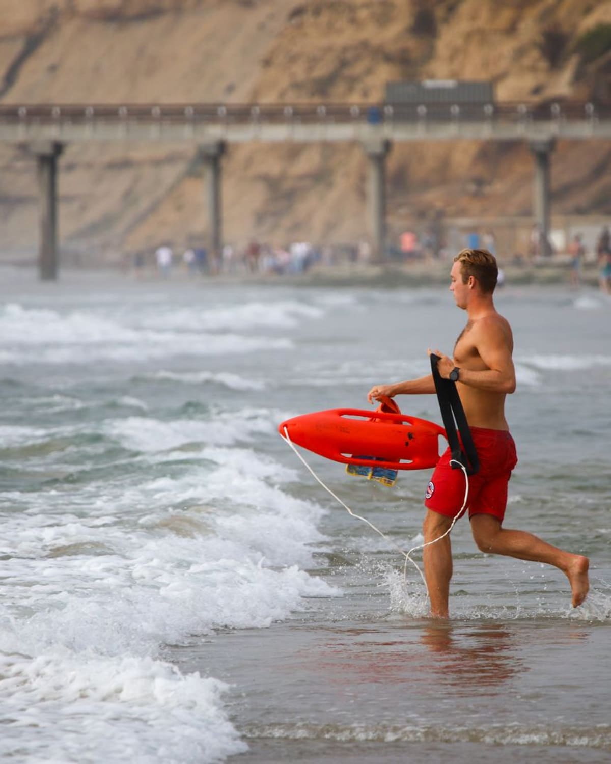 A rescue swimmer runs into the surf carrying a red rescue buoy.