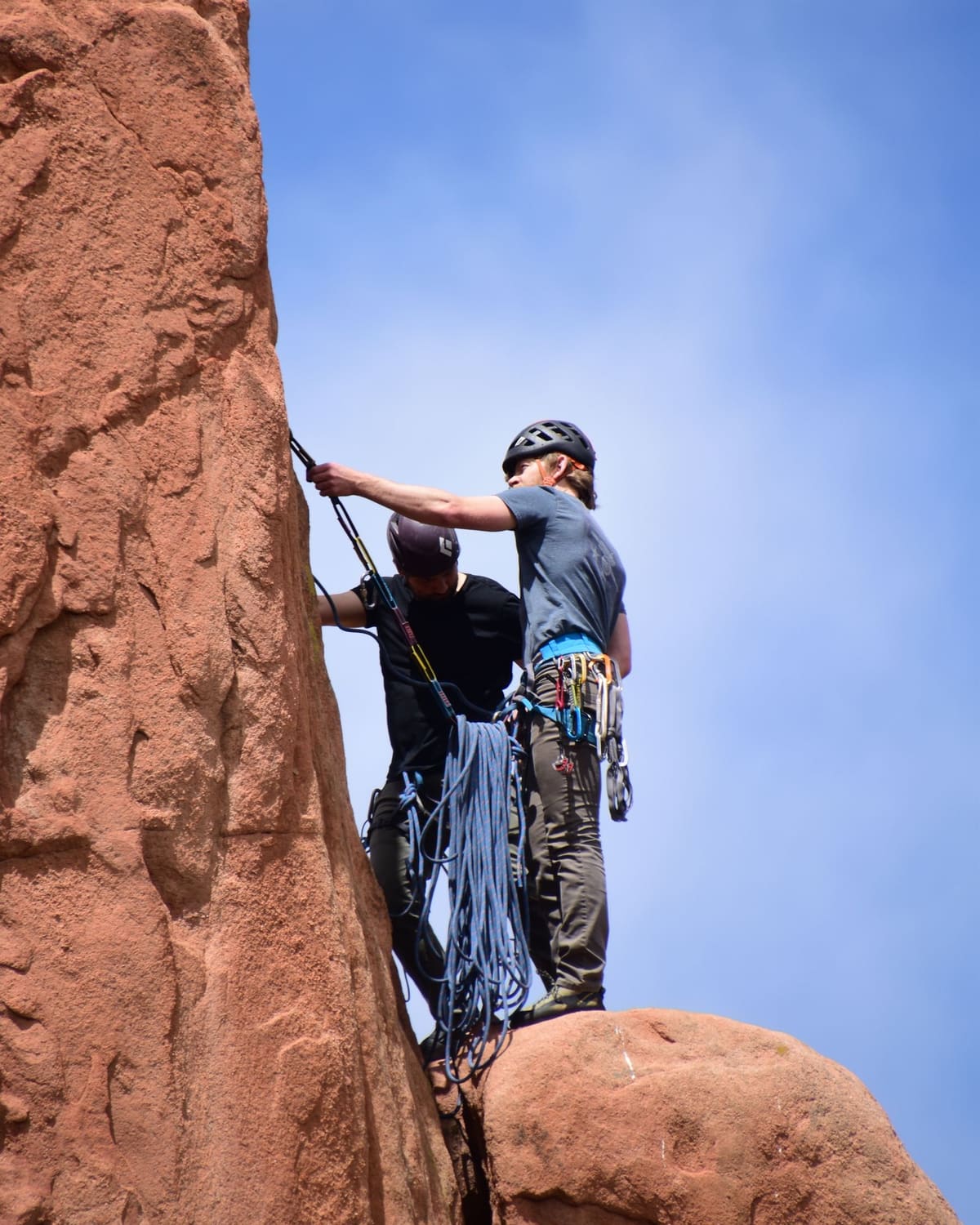Two climbers in helmets managing a rope at the top of a red sandstone cliff.