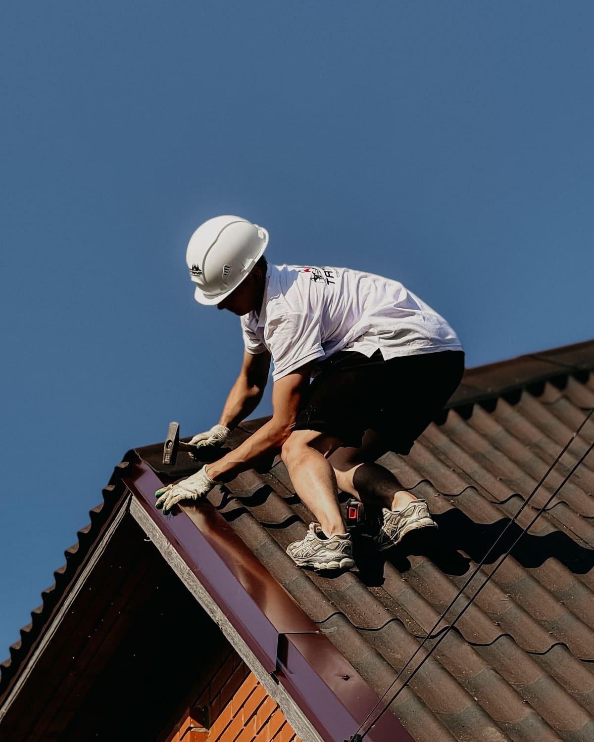 A young roofer in a white helmet crouched on a tiled roof with a hammer, working the edge against a clear blue sky.