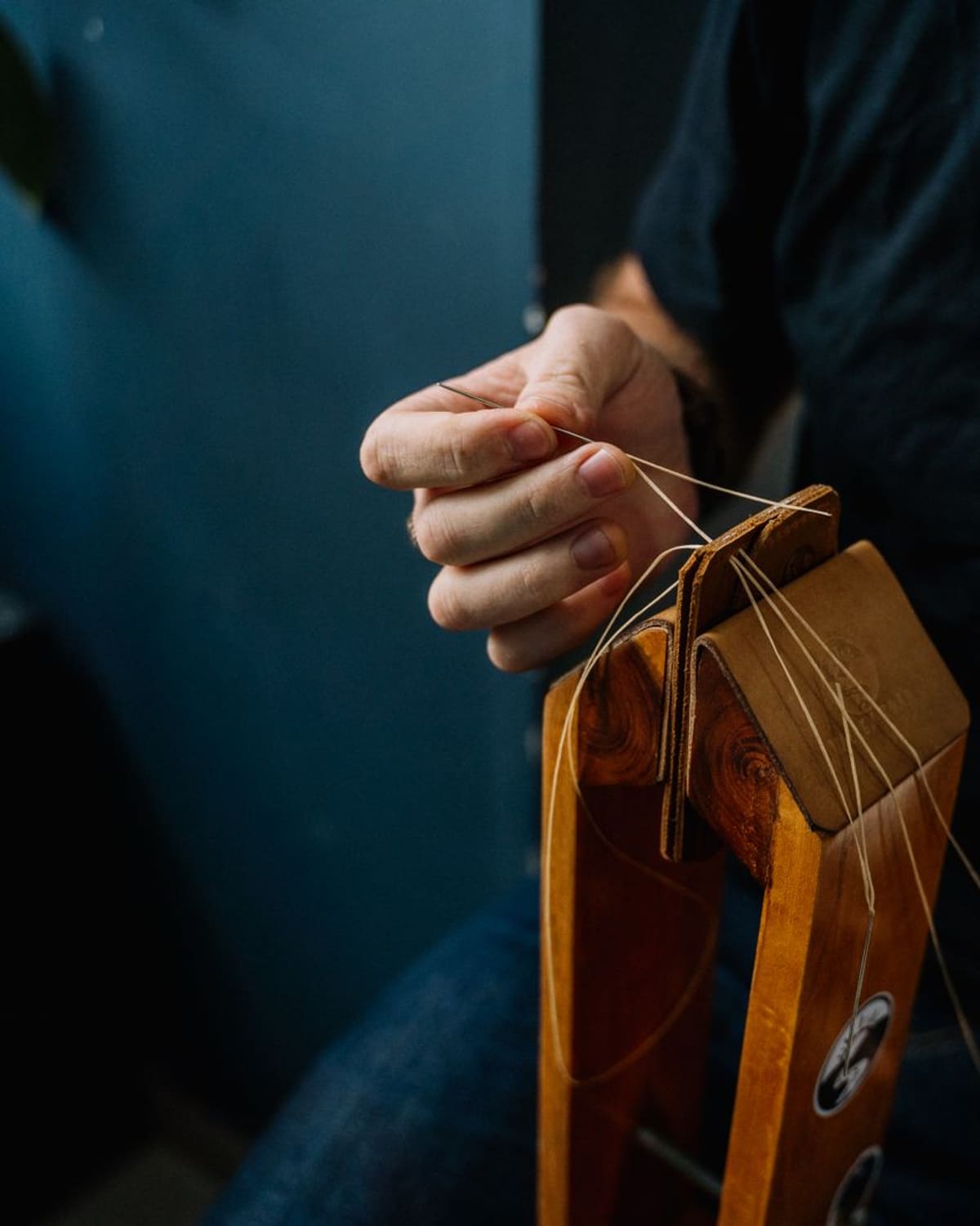 Hands hand-stitching a piece of leather held upright in a wooden stitching clamp, thread pulled taut.