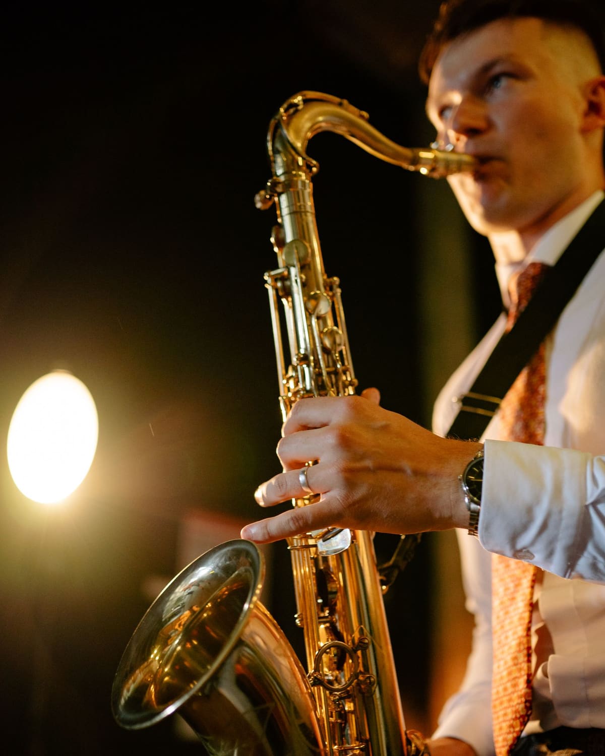 A young saxophonist in a waistcoat plays a tenor saxophone under warm stage light.