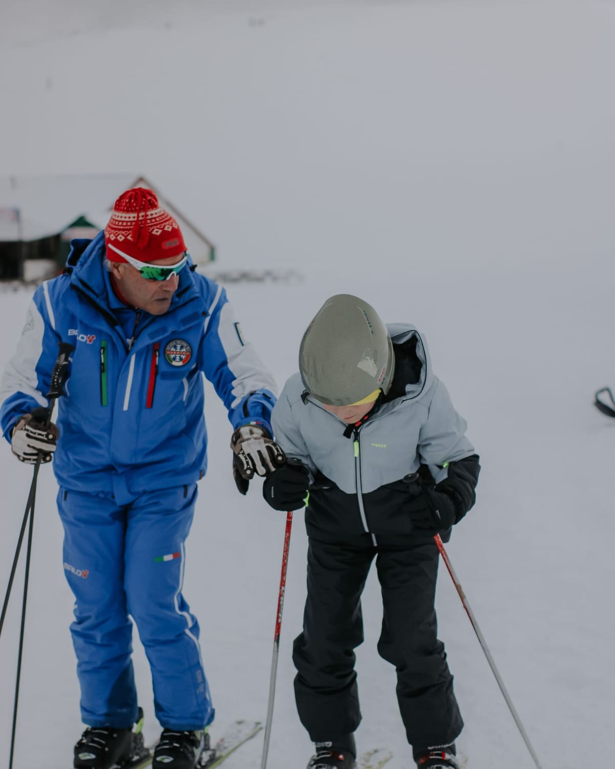 Ski instructor in a blue jacket holding a young learner's hand on a snowy beginner slope.