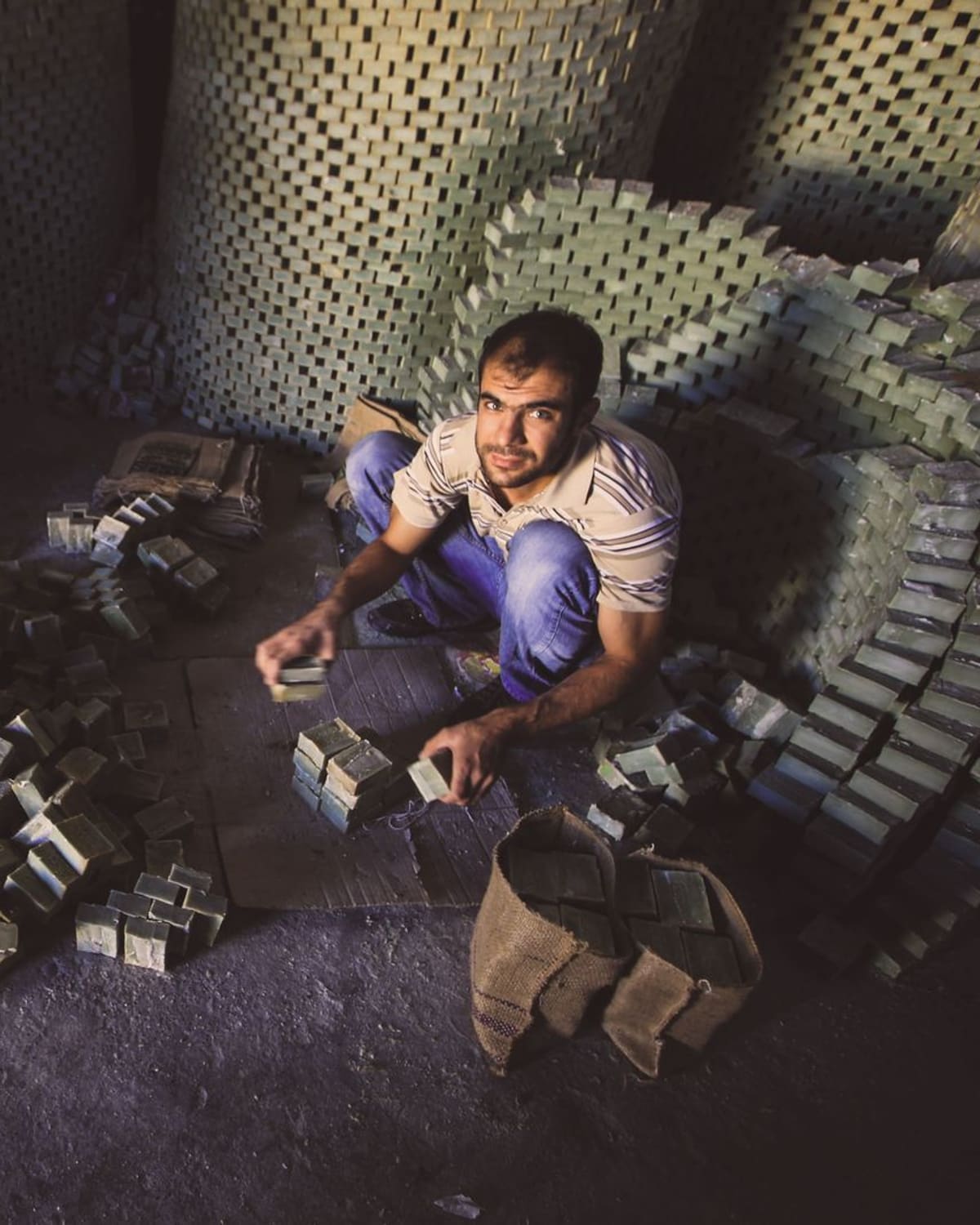 A man crouched among tall lattice towers of curing soap, arranging stamped soap blocks by hand in a dim workshop lit by a shaft of daylight.