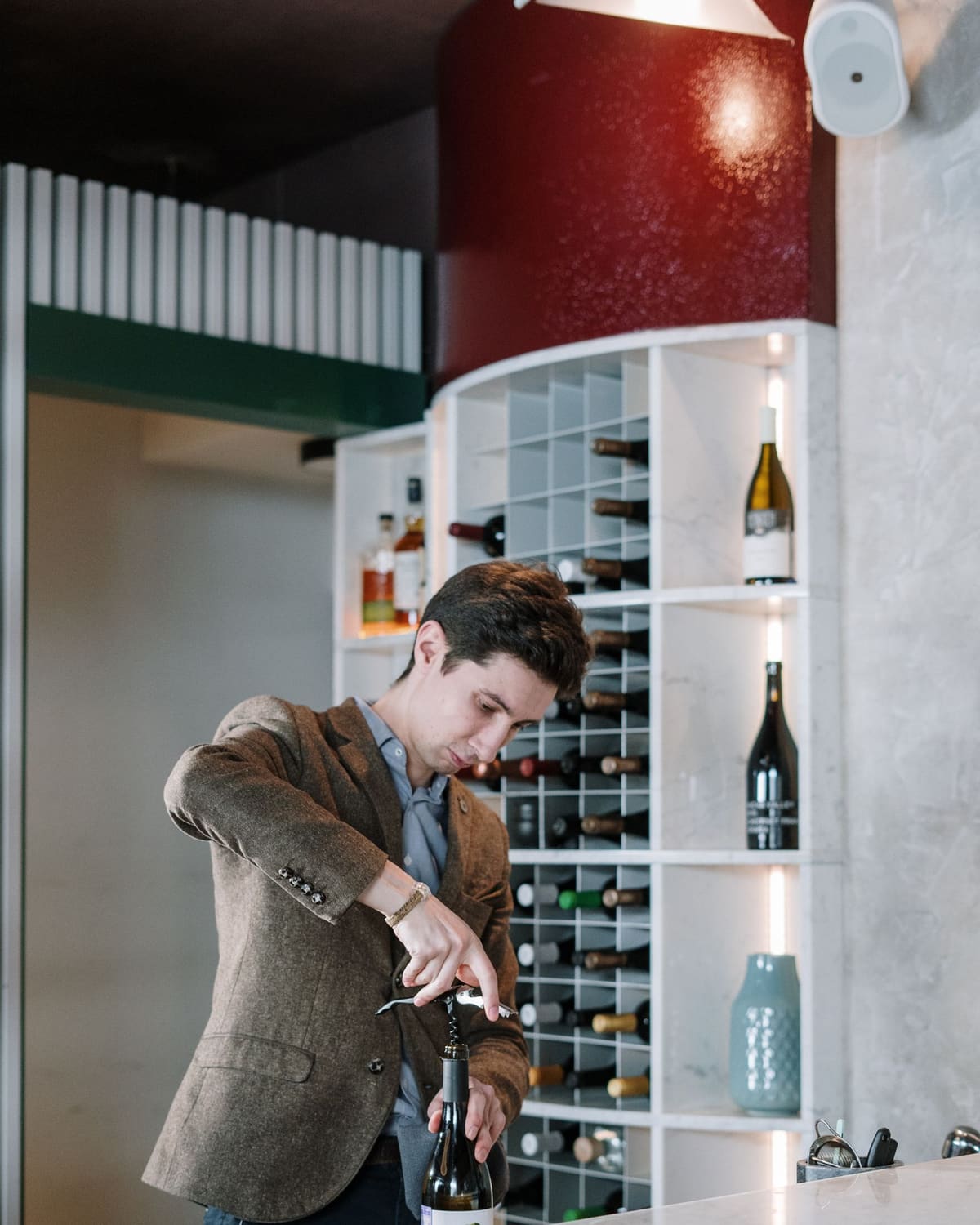 A sommelier in a tweed jacket opening a bottle before a wall of wine