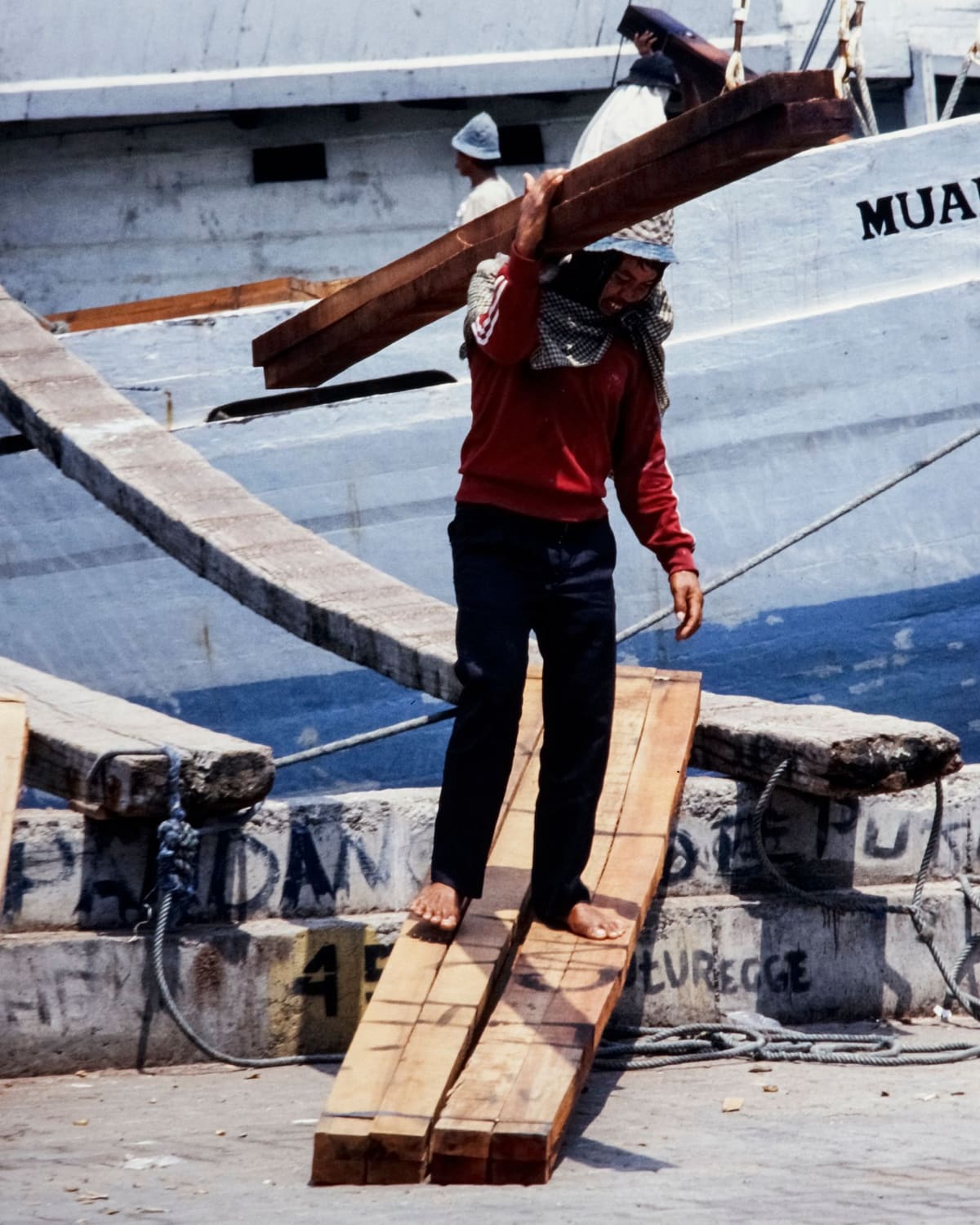 Dock worker carrying heavy wooden beams on his shoulder down a plank from a ship.