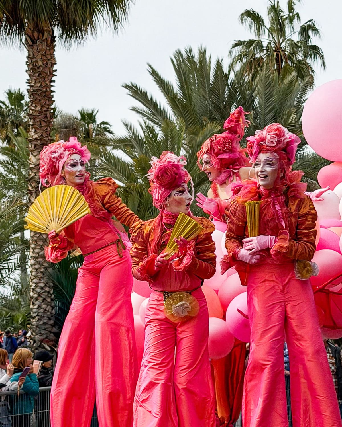 Carnival stilt walkers in pink costumes with fans tower over a festival crowd.