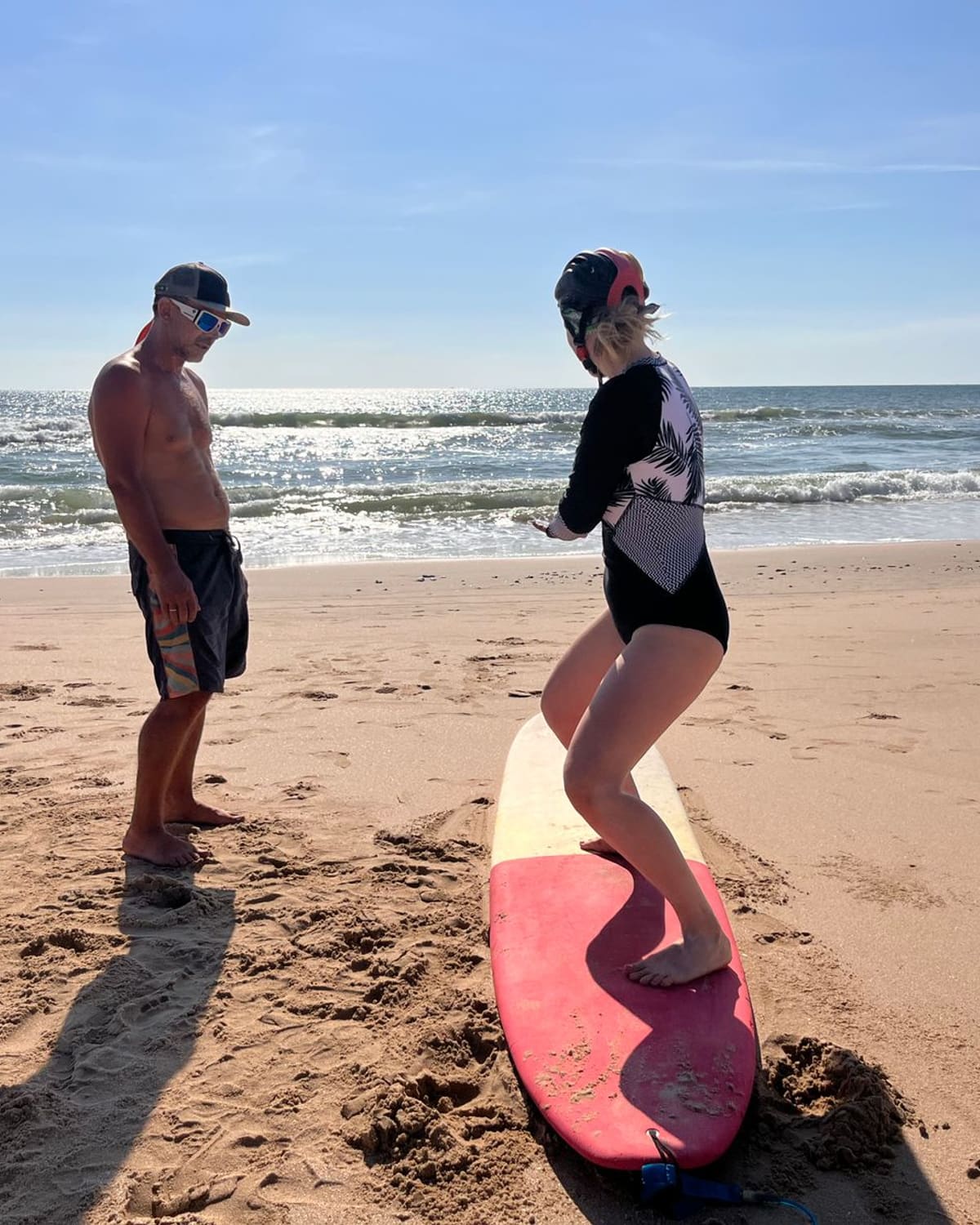 Surf instructor watching a learner practise standing on a surfboard on a sunlit beach.
