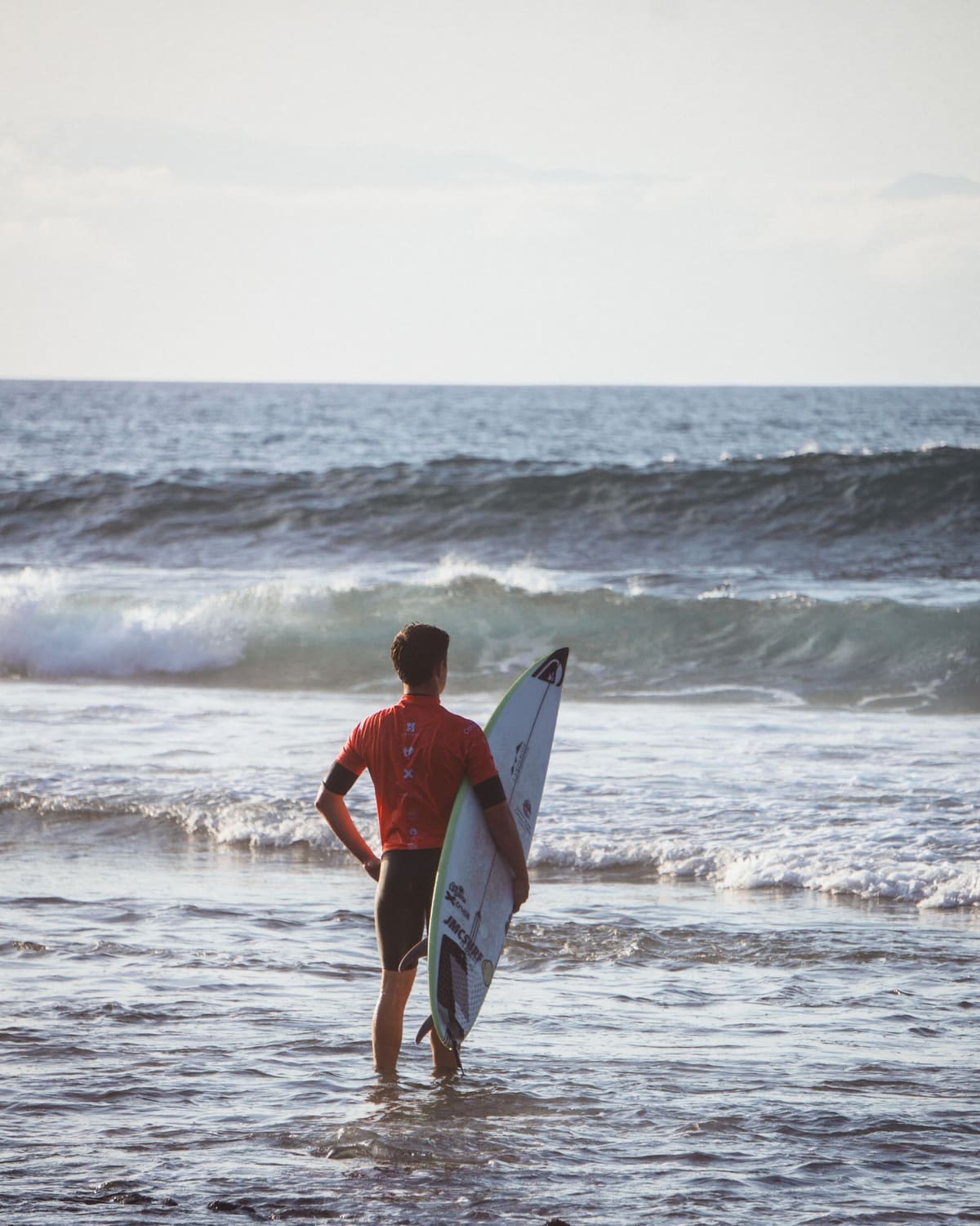 A surfer in a red top stands in the shallows at golden hour, board under one arm, reading the incoming waves.