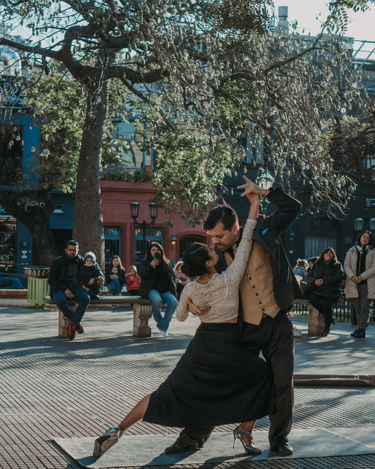 A man in a suit and a woman in a lace top lean into a tango pose on a stone plaza, with seated and standing spectators watching under bare trees in golden light.