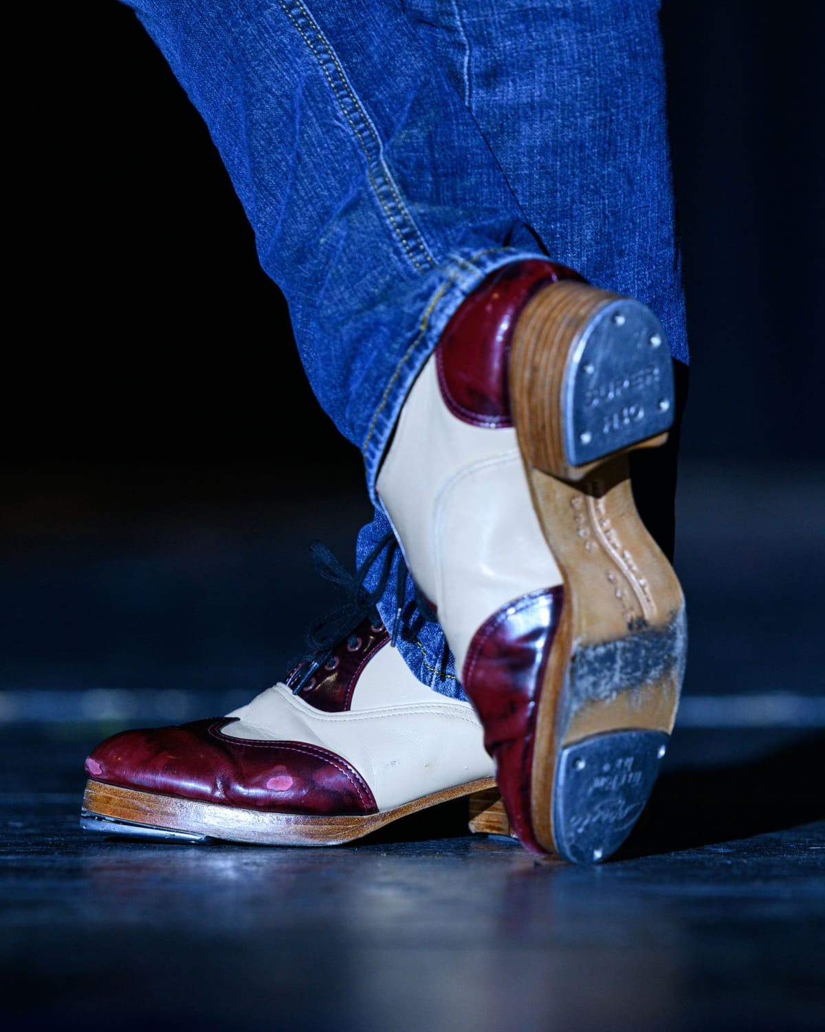 A close-up of a person's legs in jeans and vintage red-and-white tap shoes, one foot flat and one lifted to show the metal tap plate on the sole, on a dark stage floor.