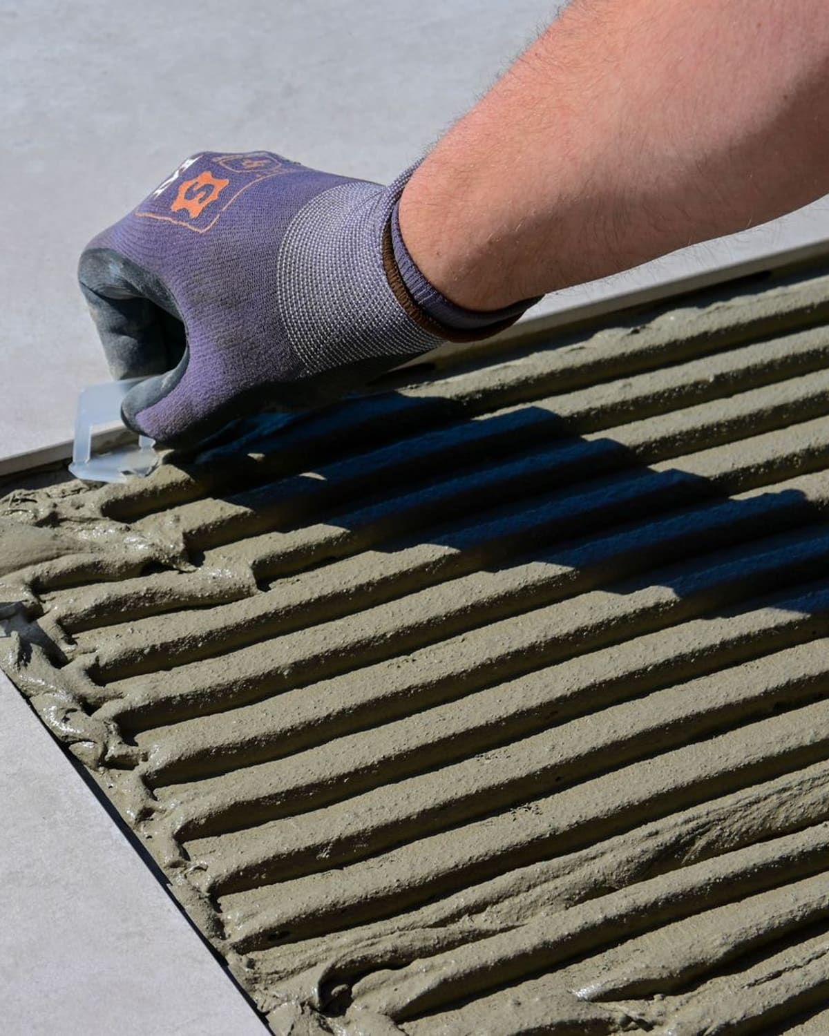 A gloved hand combing grey tile adhesive into even ridges with a notched trowel on a floor.