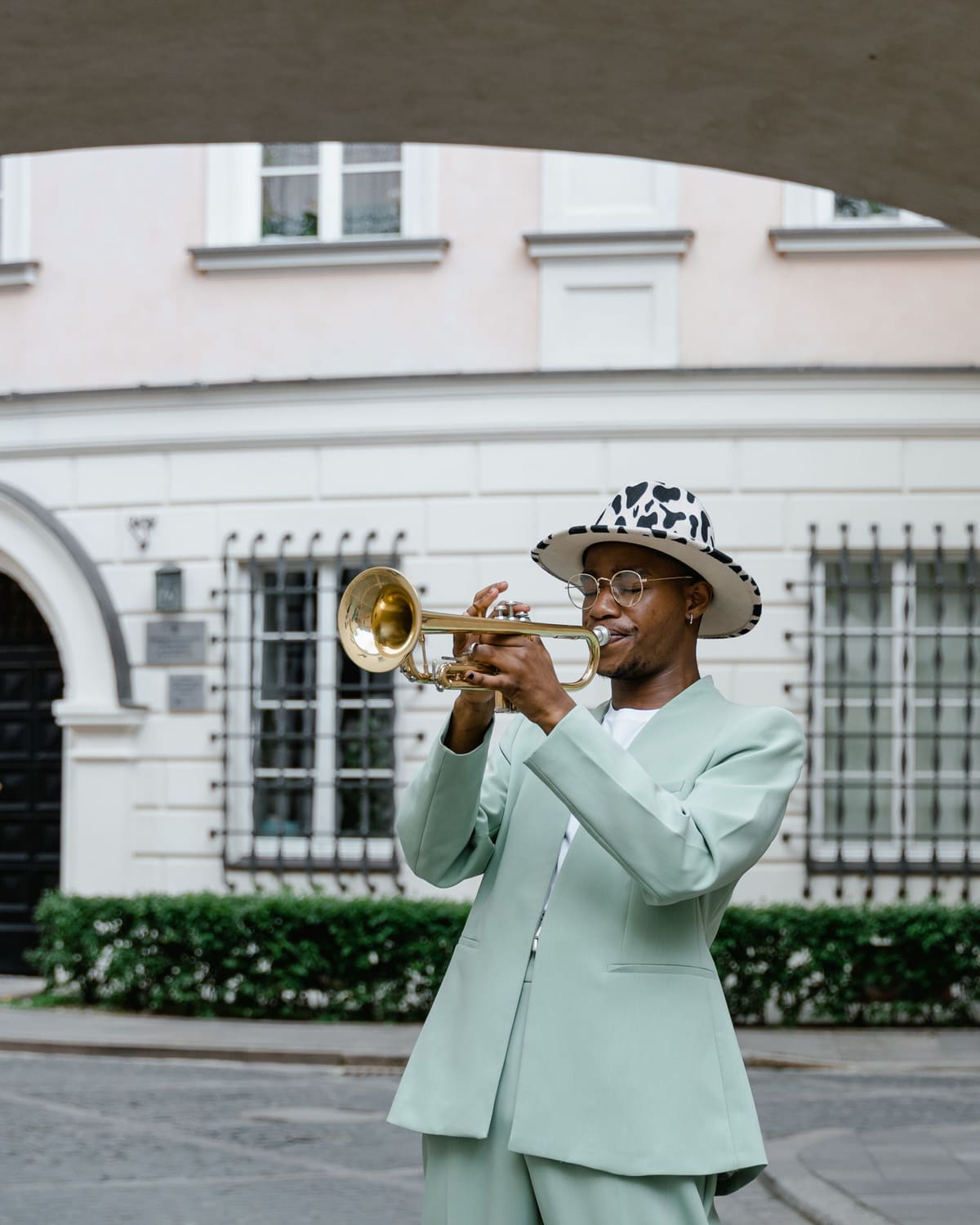 A trumpeter in a mint suit and patterned hat plays a gold trumpet on a city street.