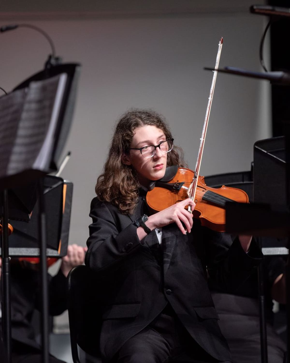 A young violinist in a black jacket plays the violin on stage, surrounded by other orchestra musicians and music stands under warm light.