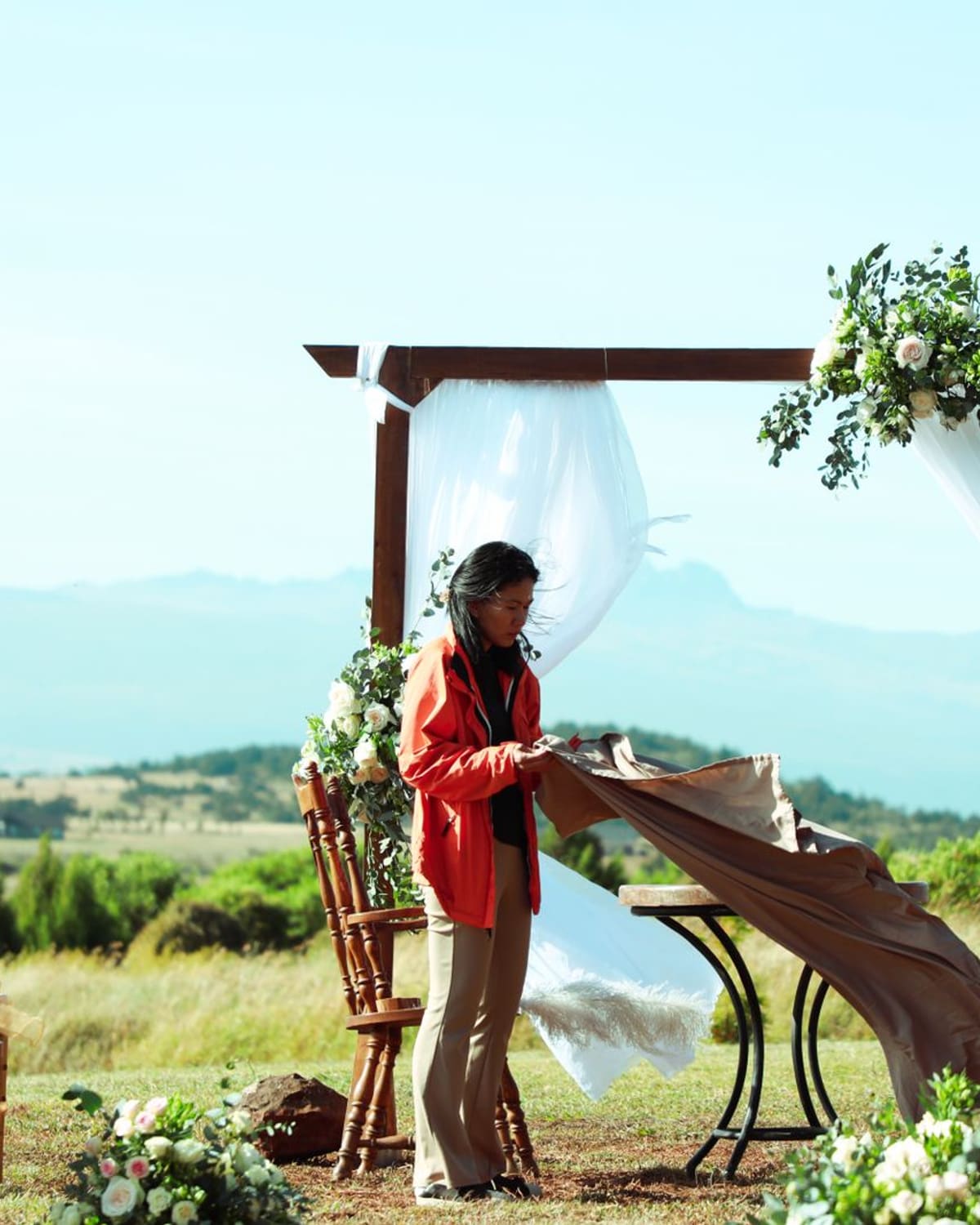 A woman in an orange jacket smooths a cloth over a table while a man adjusts a flower-covered wooden arch draped in white fabric, on a grassy hilltop with mountains behind.