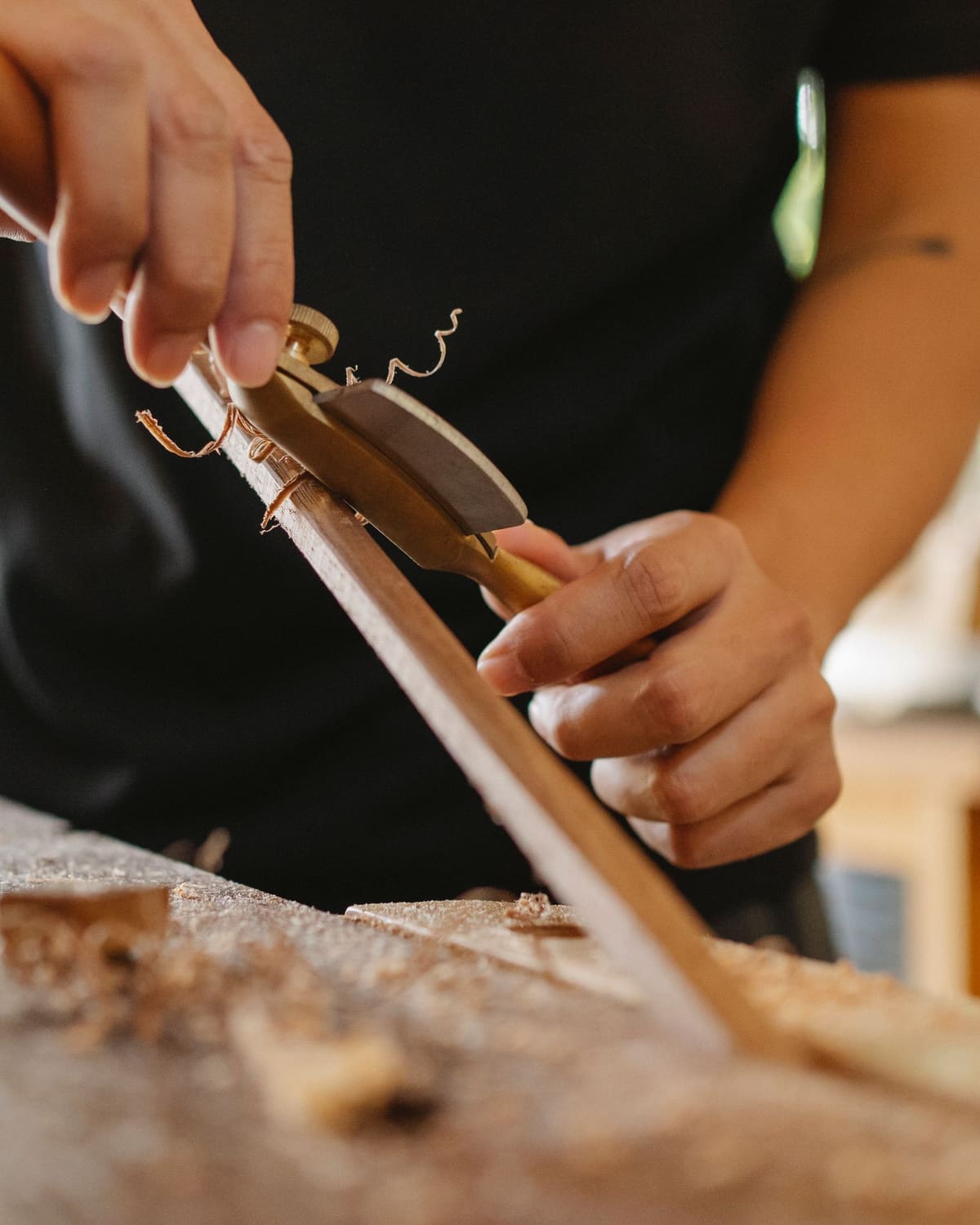 A maker's hands shaping a wooden shaft with a spokeshave, curls of shaving on the workbench