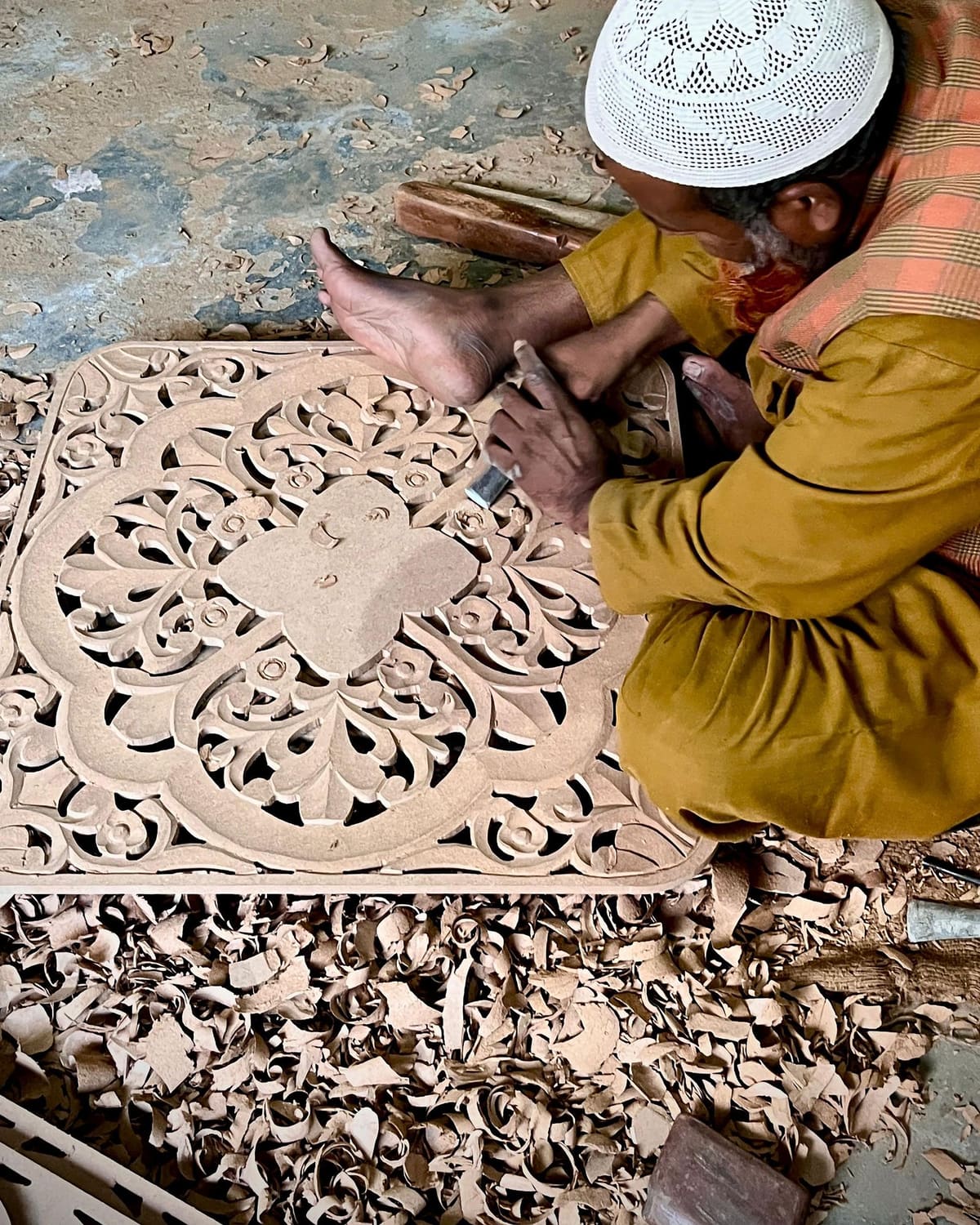 A craftsman in a white cap carves an intricate floral pattern into a wooden panel, surrounded by wood shavings.
